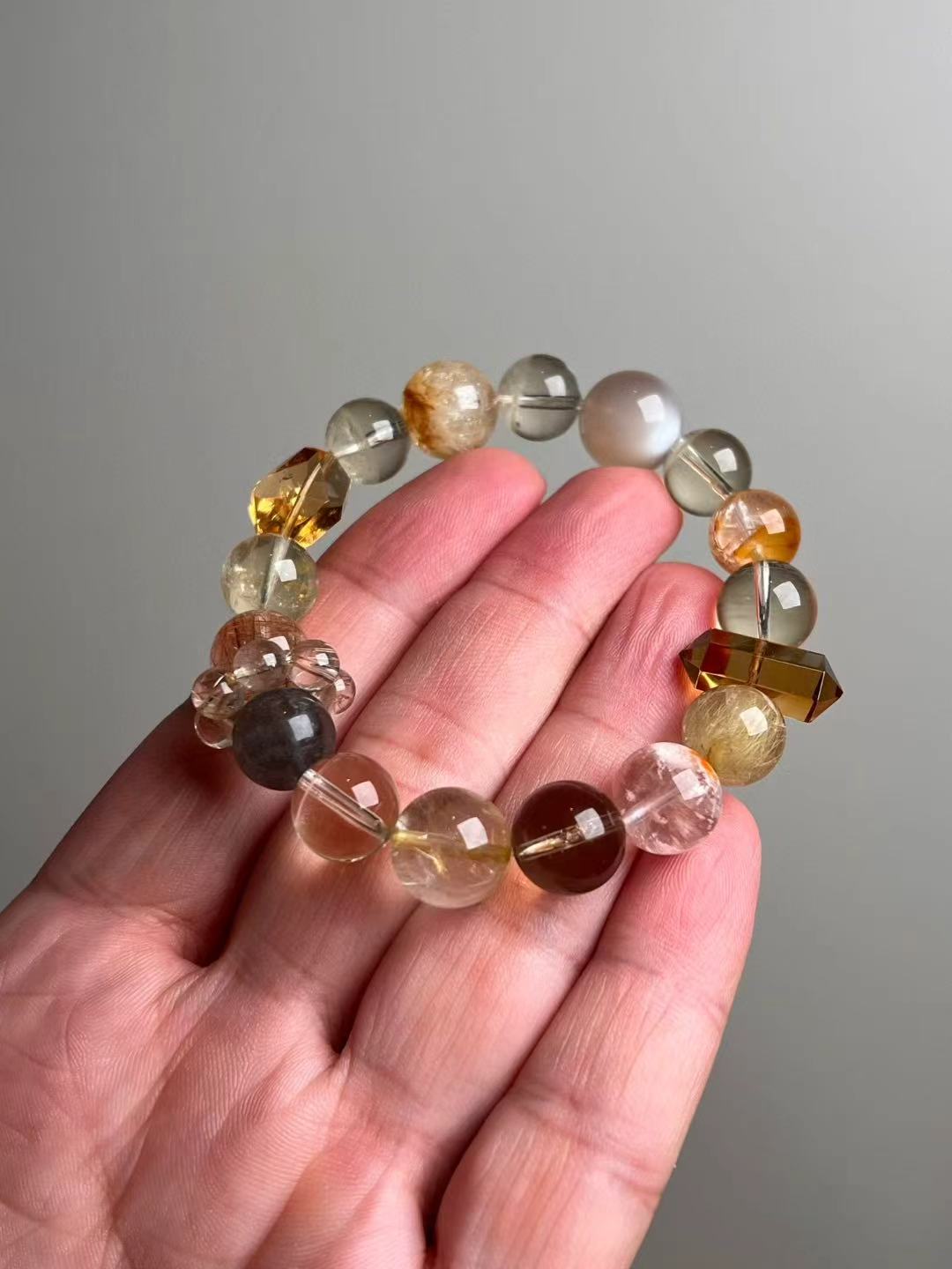 Hand closeup of gold-toned bracelet with golden rutilated quartz, citrine, grey/orange moonstone, garden quartz and a rutile runner cluster (14–11 mm)