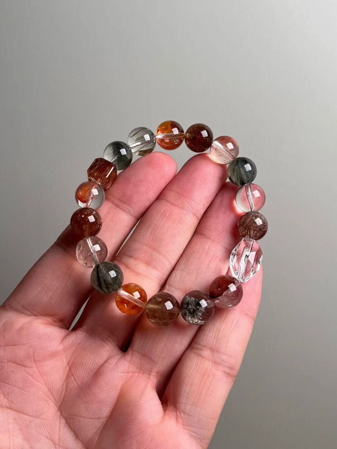Hand shot of the multigem energy bracelet—10–11 mm rounds mixing green phantom, black-gold super seven, red lepidocrocite, green rutilated quartz, ghost and clear quartz, plus Auralite-23.